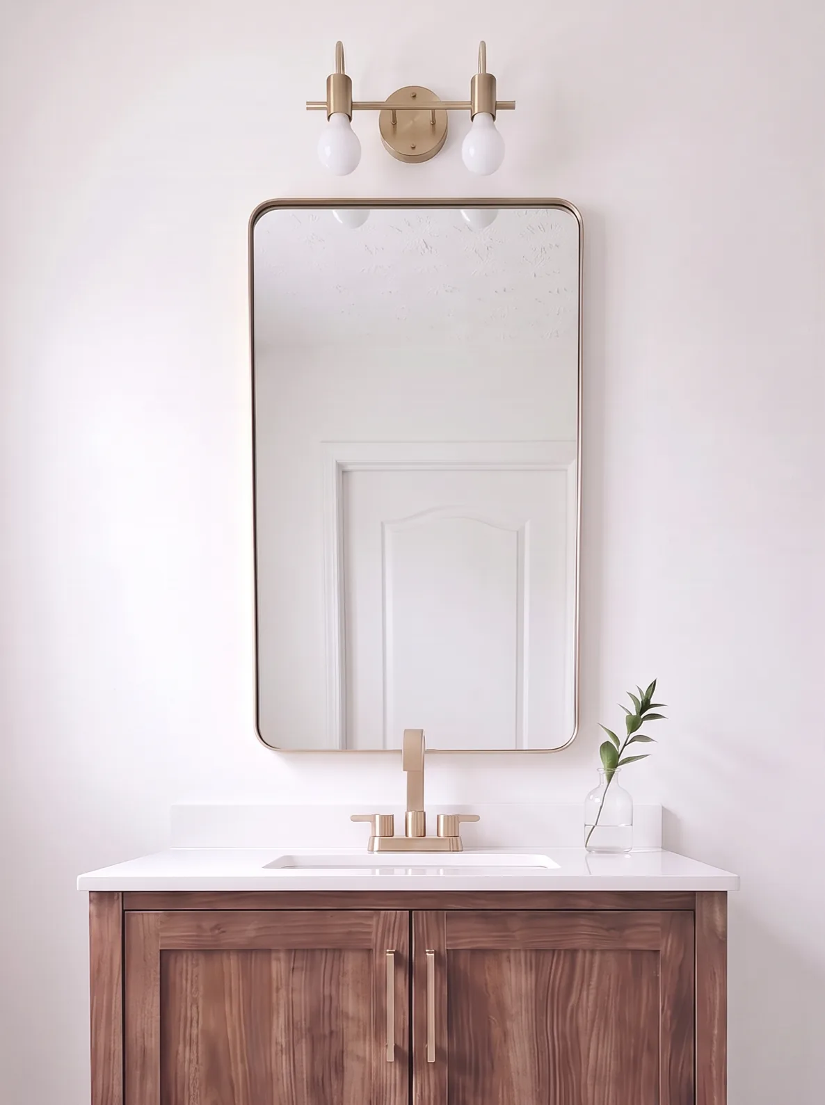 The walnut vanity straight on: brass two-light sconce, brass-framed mirror, brass faucet and white quartz top on a marble-look floor