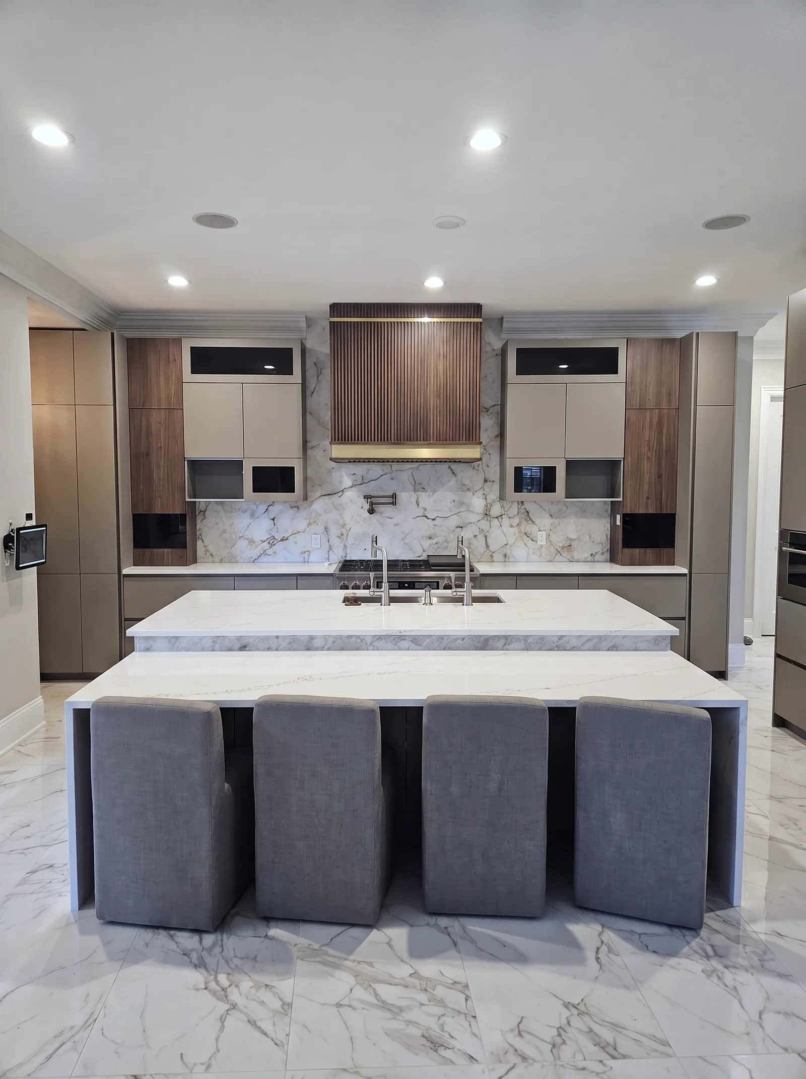 The kitchen straight on: a fluted walnut range hood centred over the range and a full-height stone slab, flat-panel cabinets either side, and a two-level white quartz island with four grey stools in front