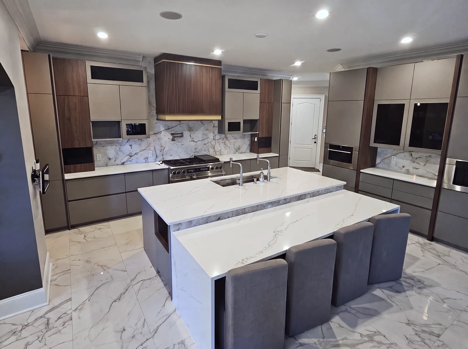 A modern kitchen with a two-level white quartz island and four grey stools, a fluted walnut range hood, greige flat-panel cabinets and a polished marble-look floor