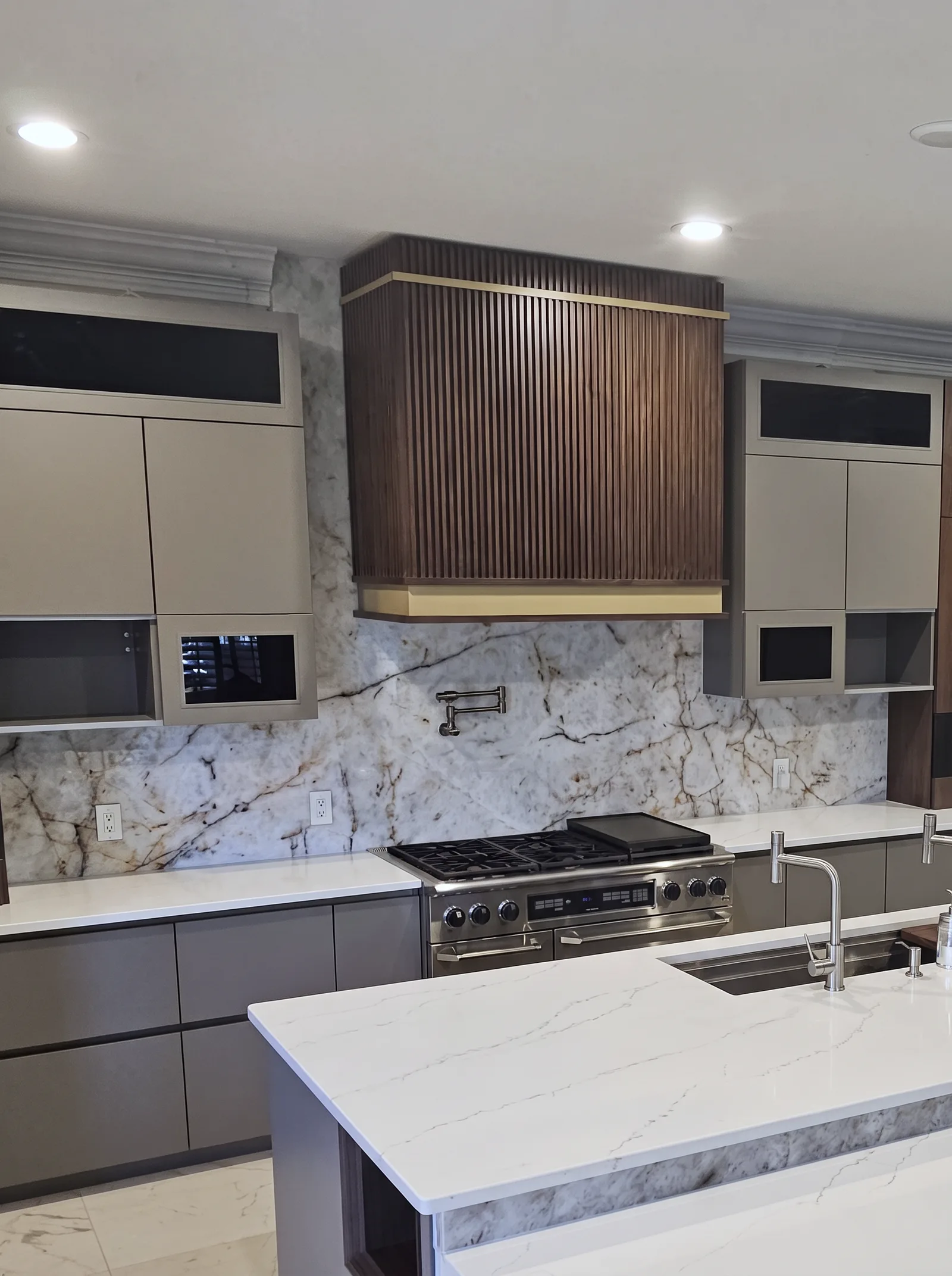 The range wall: a fluted walnut hood with a brass band over a stainless gas range, a full-height stone slab backsplash with a pot filler, and flat-panel upper cabinets