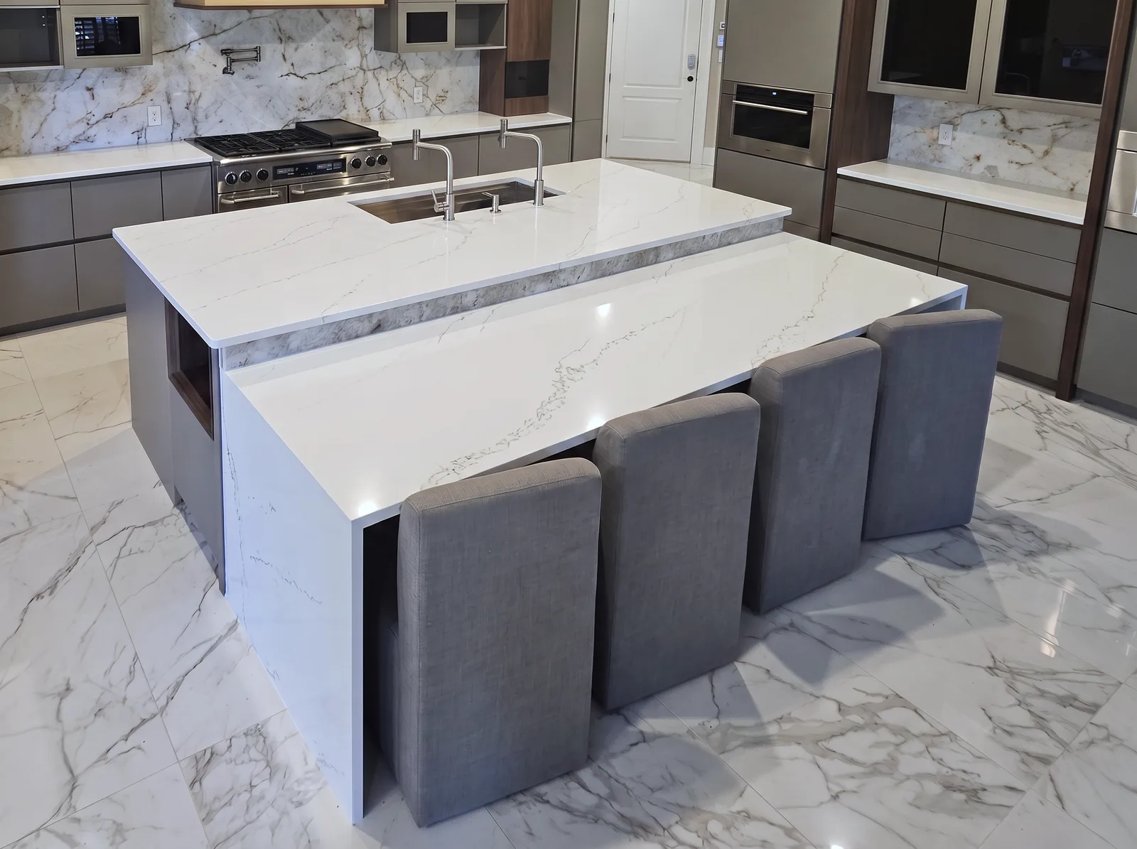 The island: a raised quartz top with a sink and two faucets, a stone-clad step and a lower waterfall counter with four grey stools