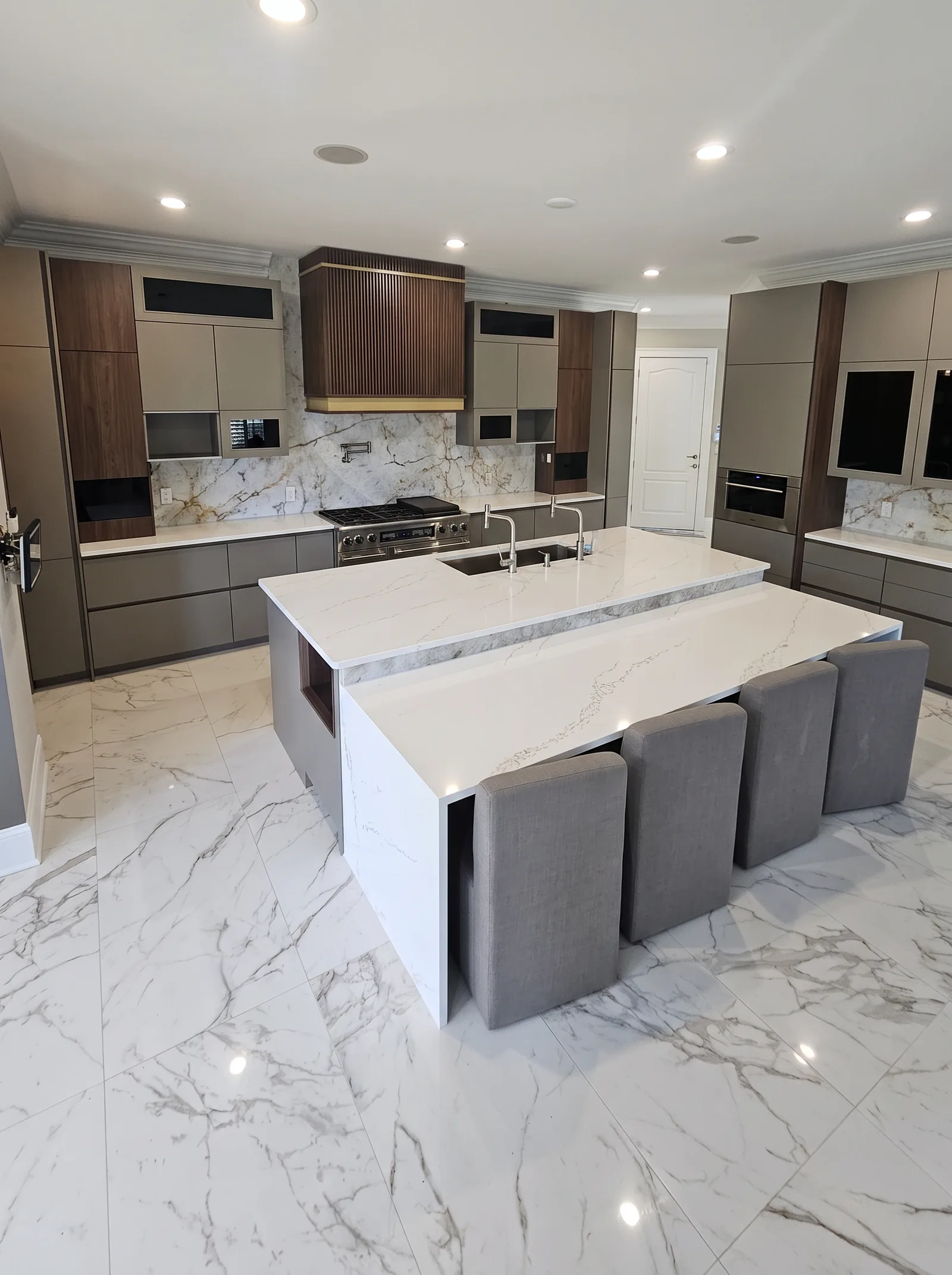 The whole kitchen from the near end: the island in front of the range wall, tall cabinets with a wall oven at the right, and the marble-look tile floor