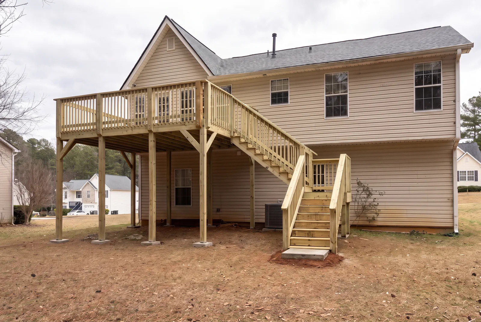 A two-storey pressure-treated pine deck on the back of a tan house, on posts with knee braces, with stairs down along the wall to a landing and the yard
