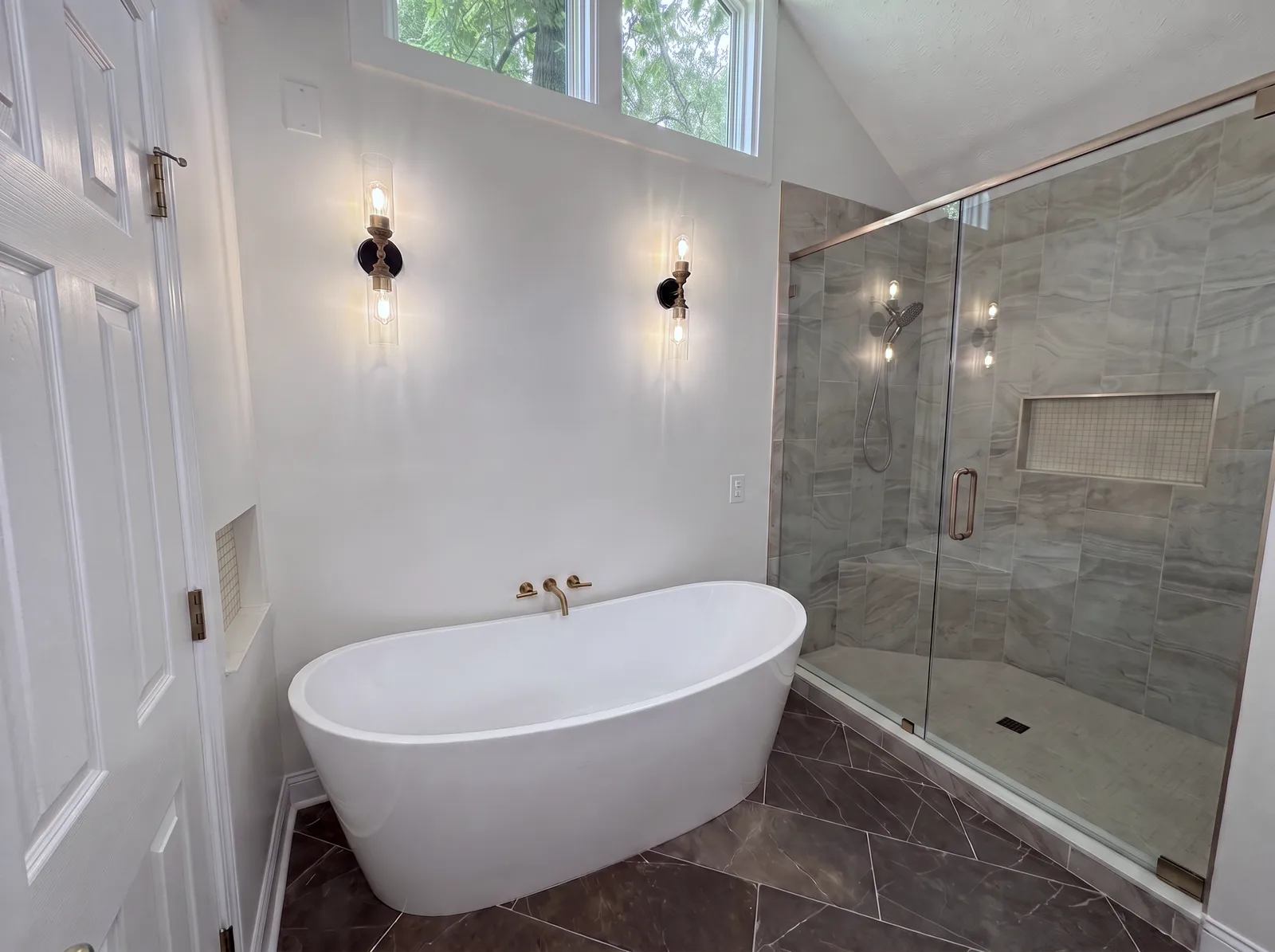 A freestanding soaking tub with a gold wall-mounted filler and two sconces, beside a walk-in shower with grey tile, a corner bench and frameless glass