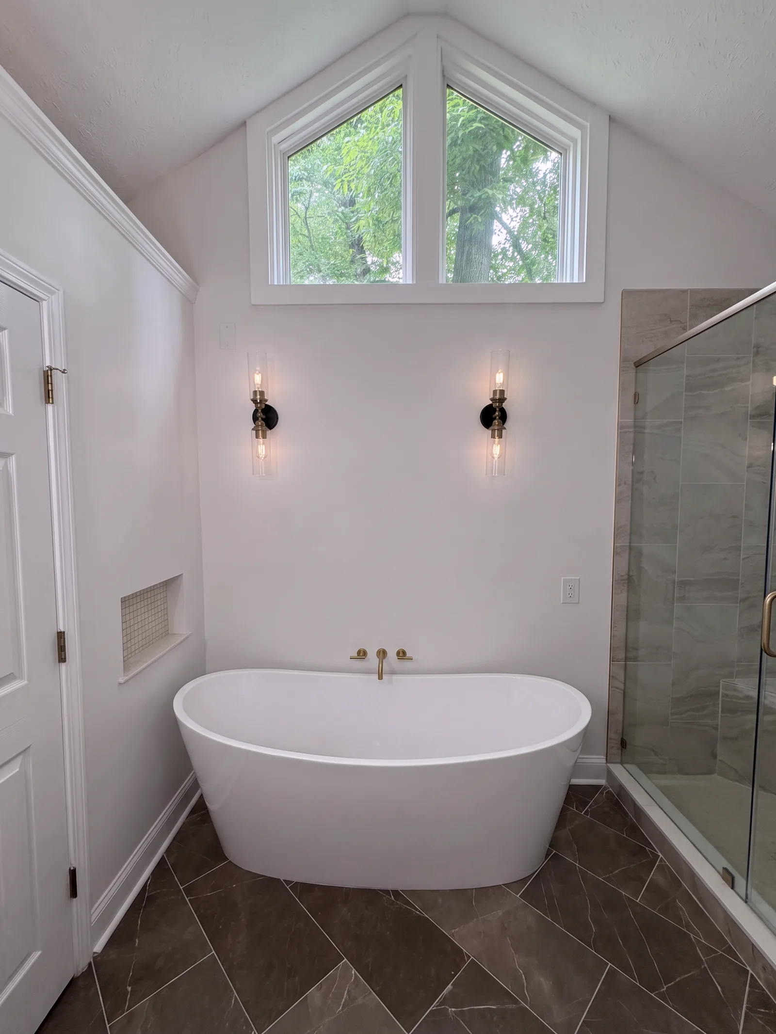 The soaking tub centred under a pair of gable windows, with a small tiled niche in the left wall and a dark herringbone tile floor