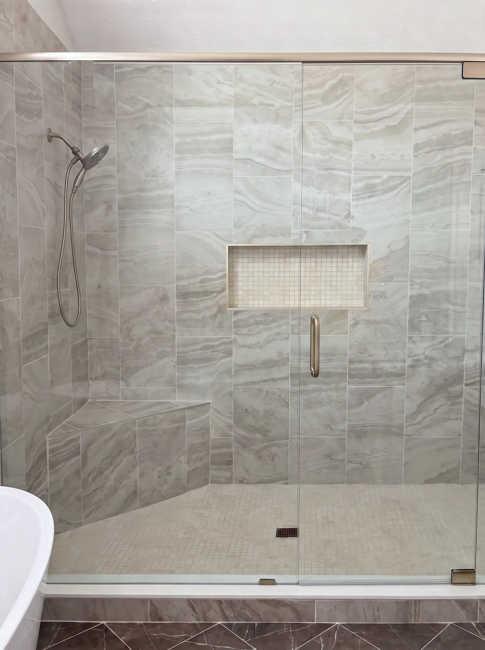 The walk-in shower through its glass: upright grey stone-look tile, a corner bench, a recessed niche with a mosaic back and a mosaic floor