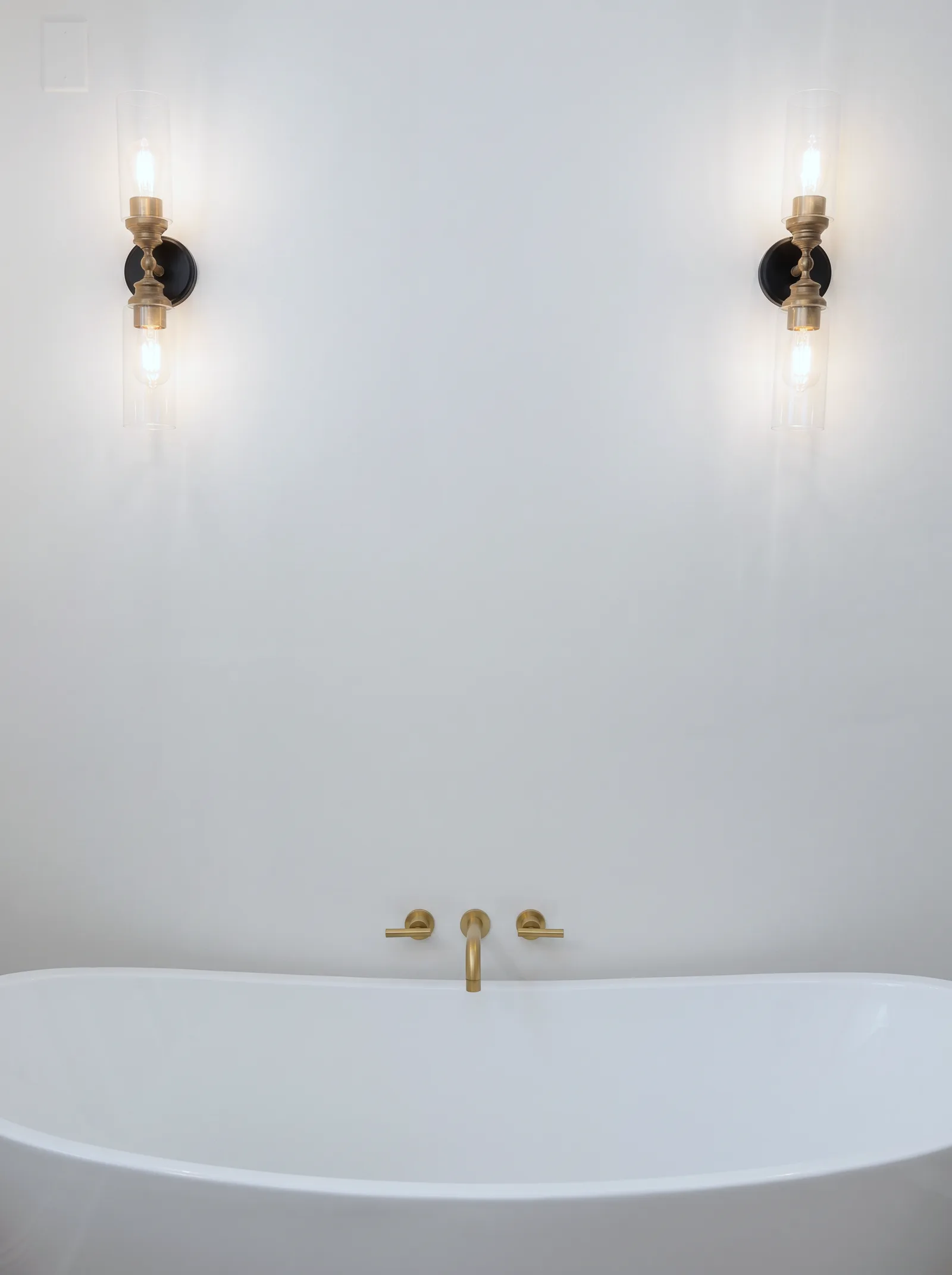 Detail of the tub wall: two black and brass sconces with clear glass shades above a brushed gold tub filler